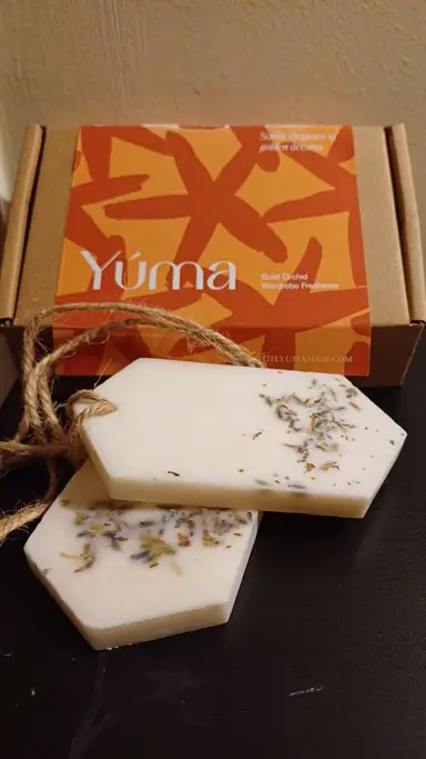 Δύο λευκά αρωματικά κεριά ντουλάπας Gold Orchid της YUMA με αποξηραμένα άνθη μπροστά από τη συσκευασία τους.