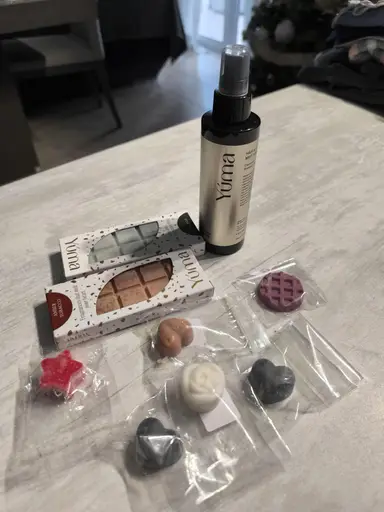 Ένα σπρέι YUMA δίπλα σε δύο συσκευασίες wax melts και διάφορα αρωματικά κεριά σε σχήματα.