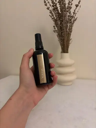 Ένα χέρι κρατά το Eternal Eden room spray της YUMA μπροστά από βάζο με αποξηραμένη λεβάντα.