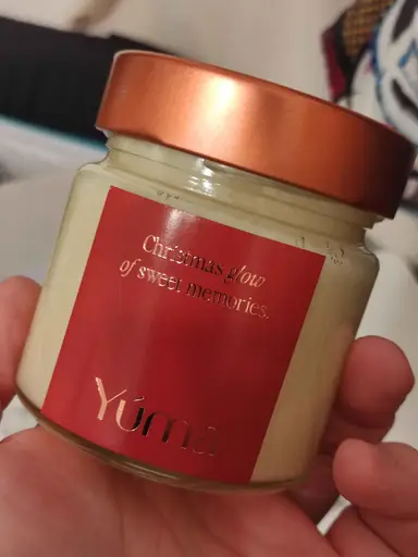 Κερί της YUMA σε γυάλινο βαζάκι με χάλκινο καπάκι και κόκκινη ετικέτα που κρατιέται στο χέρι.