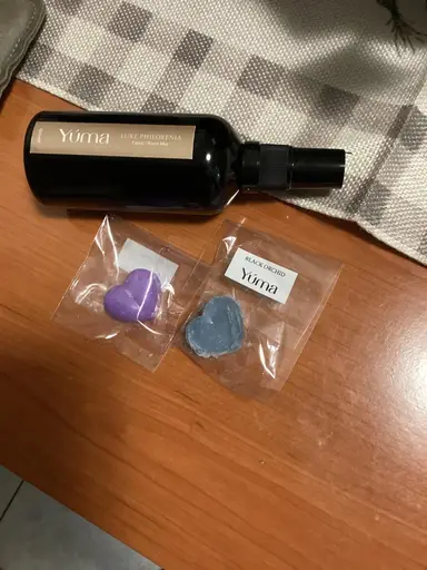 Ένα μαύρο μπουκάλι room spray YUMA δίπλα σε δύο αρωματικά κεριά σε σχήμα καρδιάς.