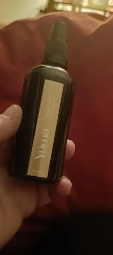 Ένα χέρι κρατά ένα σκουρόχρωμο μπουκάλι room spray της YUMA με χρυσή ετικέτα.