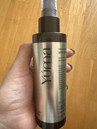 Ένα χέρι κρατάει ένα κυλινδρικό μπουκάλι Hair & Body Mist της YUMA με ασημί ετικέτα.
