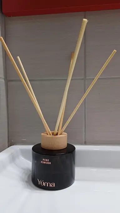 Μαύρο γυάλινο reed diffuser της YUMA με ξύλινο πώμα και πέντε ξυλάκια μπροστά από γκρι πλακάκια.