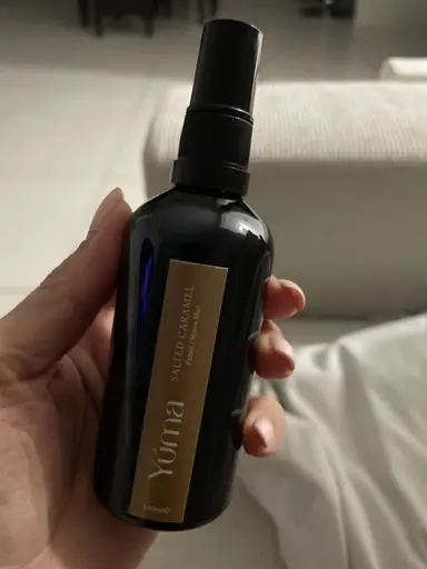 Ένα χέρι κρατά το Salted Caramel Room Mist της YUMA σε μαύρο γυάλινο μπουκάλι με χρυσή ετικέτα.