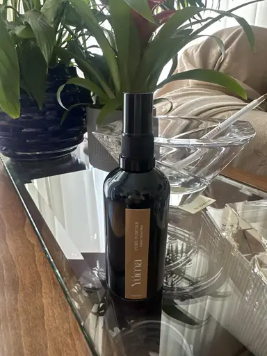 Ένα μαύρο μπουκάλι room spray της YUMA πάνω σε γυάλινο τραπέζι δίπλα σε πράσινα φυτά εσωτερικού χώρου.