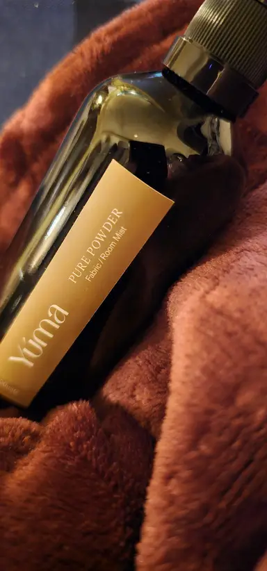 Σκούρο γυάλινο μπουκάλι Room Mist Pure Powder της YUMA τοποθετημένο πάνω σε μαλακό καφέ ύφασμα.