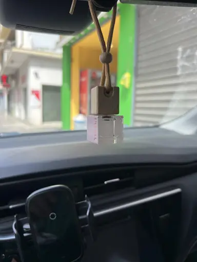 Ένα μικρό γυάλινο μπουκαλάκι αρωματικού αυτοκινήτου της YUMA με ξύλινο πώμα κρέμεται από τον καθρέφτη.