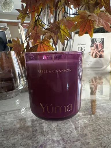 Μωβ αρωματικό κερί YUMA Apple & Cinnamon σε γυάλινο δοχείο μπροστά από φθινοπωρινά φύλλα.