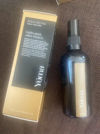 Ένα σπρέι χώρου Gold Orchid της YUMA δίπλα στη χρυσή και μαύρη συσκευασία του.