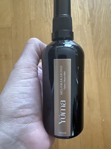 Ένα χέρι κρατά το μαύρο μπουκάλι Room Mist Melomakarono της YUMA μπροστά από ξύλινο δάπεδο.