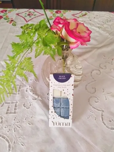 Συσκευασία Black Orchid της YUMA δίπλα σε βάζο με τριαντάφυλλα πάνω σε κεντημένο λευκό τραπεζομάντηλο.