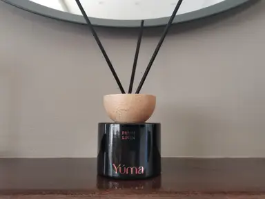 Μαύρο γυάλινο αρωματικό χώρου YUMA Fresh Linen με ξύλινο πώμα και μαύρα στικς σε ξύλινη επιφάνεια.