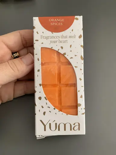 Ένα χέρι κρατάει τη λευκή συσκευασία Orange Spices της YUMA με πορτοκαλί wax melts.
