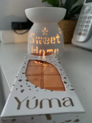 Πορτοκαλί wax melt της YUMA σε λευκή συσκευασία μπροστά από αναμμένο κεραμικό αρωματιστή χώρου.