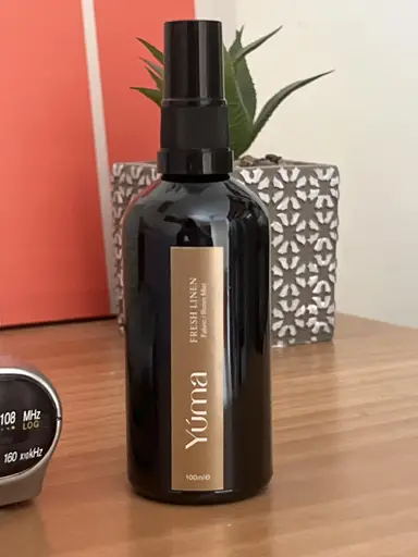 Μαύρο μπουκάλι room spray Yuma Fresh Linen σε ξύλινη επιφάνεια μπροστά από διακοσμητικό φυτό.