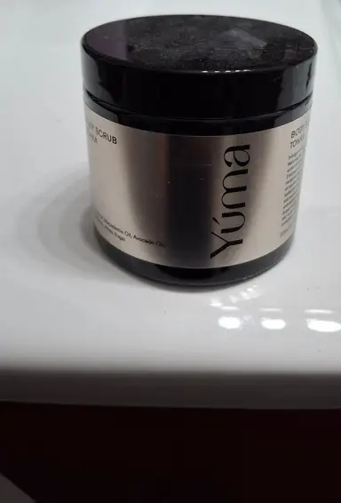 Μαύρο βάζο body scrub της YUMA με ασημί ετικέτα τοποθετημένο πάνω σε λευκή επιφάνεια.