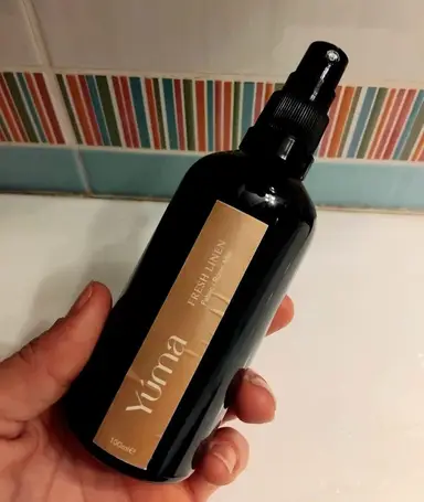 Ένα χέρι κρατά το Fresh Linen room spray της YUMA σε σκούρο μπουκάλι με μπεζ ετικέτα.