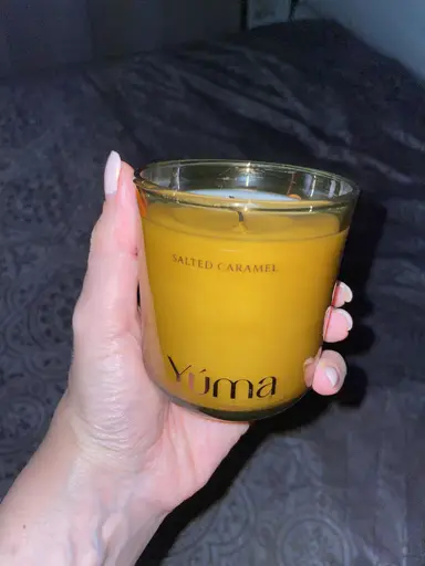 Ένα χέρι κρατά το αρωματικό κερί Salted Caramel της YUMA σε γυάλινο βαζάκι με κίτρινο κερί.