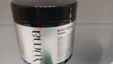 Σκούρο βάζο YUMA Body Scrub TONKA με ασημί ετικέτα και μαύρο καπάκι σε γκρίζο φόντο.