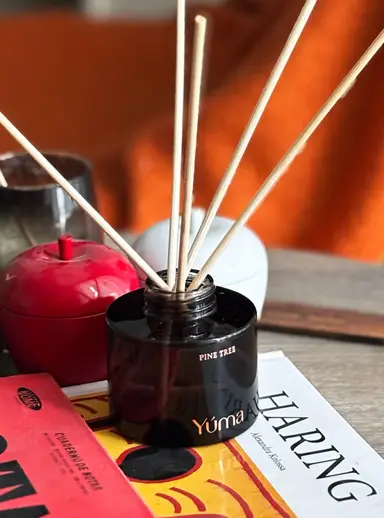Σκούρο γυάλινο δοχείο Pine Tree της YUMA με ξύλινα στικς αρωματικού χώρου πάνω σε βιβλία.