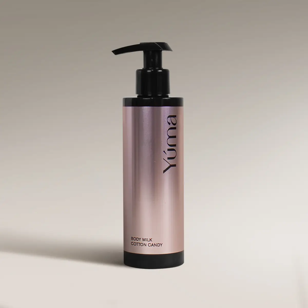 Μεταλλικό ροζ μπουκάλι Body Milk Cotton Candy της YUMA με μαύρη αντλία σε μπεζ φόντο.