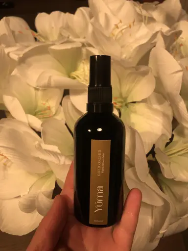 Ένα χέρι κρατά το Gold Orchid room mist της YUMA μπροστά από λευκά άνθη κρίνων.