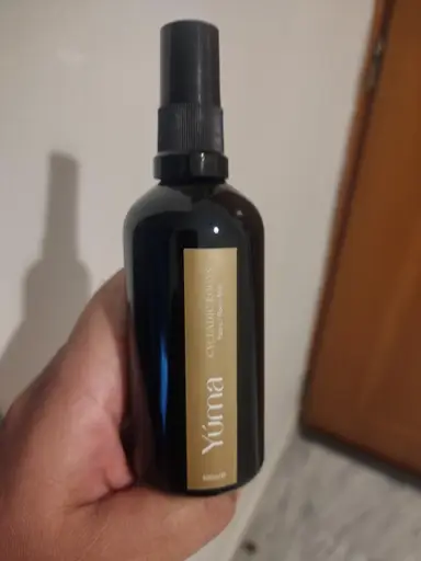 Ένα χέρι κρατά το Cycladic Roots Room Mist της YUMA σε μαύρο μπουκάλι με χρυσή ετικέτα.