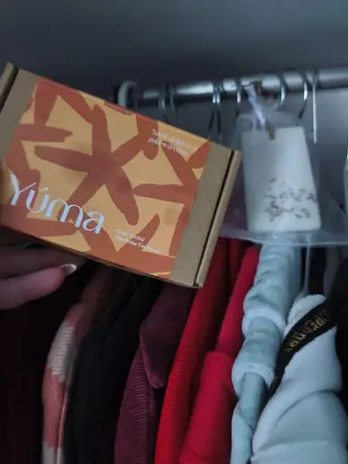 Χέρι κρατά κουτί αρωματικού ντουλάπας Gold Orchid της YUMA μπροστά από κρεμασμένα ρούχα.