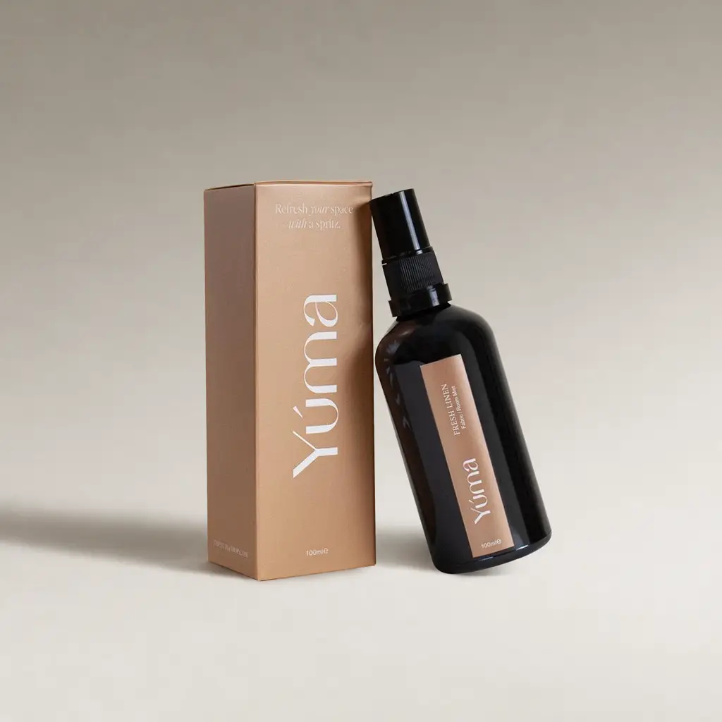 Σκούρο γυάλινο μπουκάλι Fabric Mist της YUMA δίπλα στην μπεζ χάρτινη συσκευασία του.
