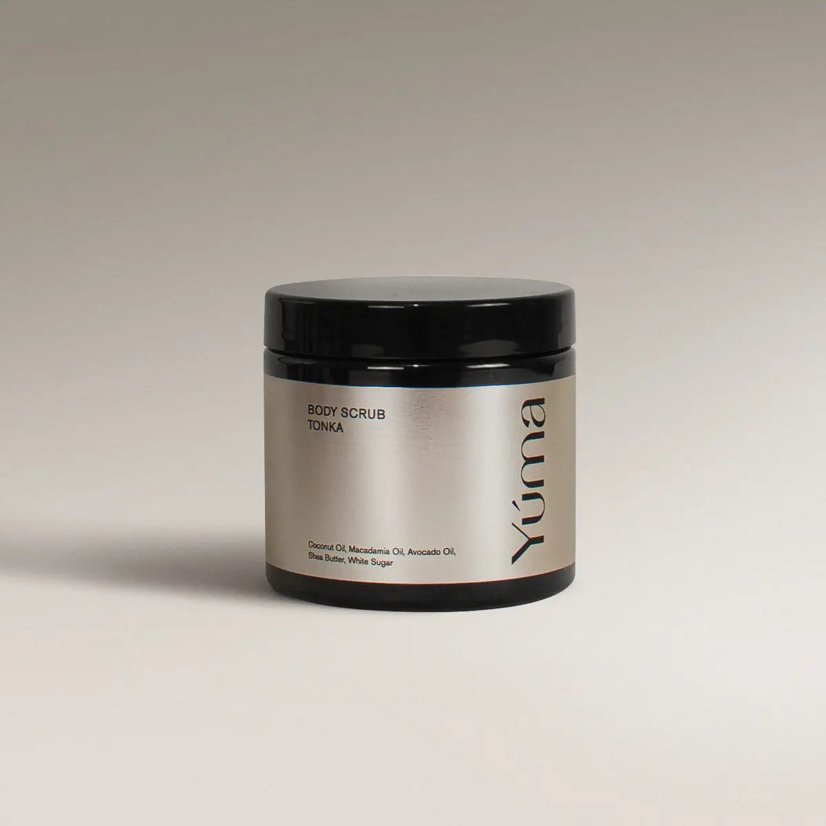 Μαύρο βάζο Body Scrub της YUMA με άρωμα Tonka και ασημί ετικέτα σε γκρίζο φόντο.
