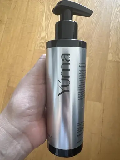 Ένα χέρι κρατάει ένα ασημί μπουκάλι της YUMA με μαύρη αντλία πάνω από ξύλινο δάπεδο.