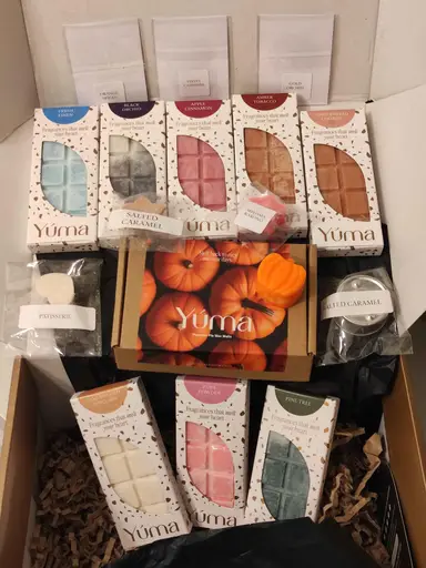 Ποικιλία από wax melts της YUMA σε διάφορα χρώματα και συσκευασίες μέσα σε χάρτινο κουτί.