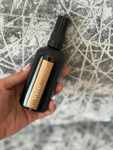 Ένα χέρι κρατά το Laundry Nostalgia room spray της YUMA μπροστά από γκρι υφασμάτινο φόντο.