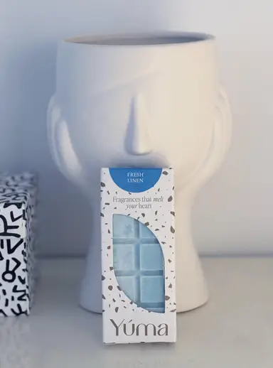 Συσκευασία wax melt Fresh Linen της YUMA μπροστά από λευκό κεραμικό βάζο σε σχήμα προσώπου.