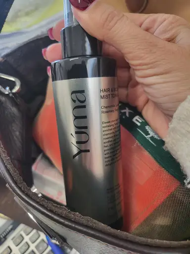 Γυναικείο χέρι κρατά ένα ασημί μπουκάλι YUMA Hair & Body Mist μέσα σε δερμάτινη τσάντα.