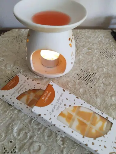 Λευκός κεραμικός καυστήρας με αναμμένο ρεσό και λιωμένο κερί δίπλα σε συσκευασίες Salted Caramel wax melts.