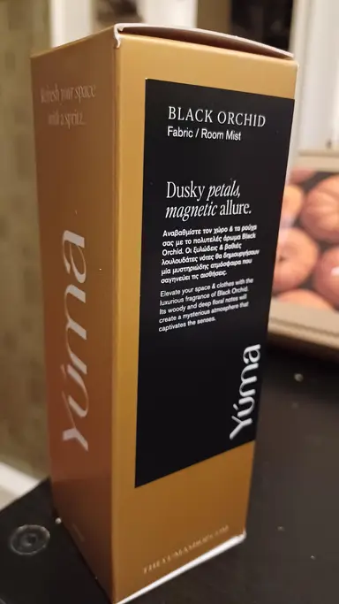 Χρυσή συσκευασία Black Orchid Fabric Room Mist της YUMA με μαύρη ετικέτα και λευκά γράμματα.