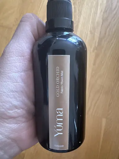 Ένα χέρι κρατά το μαύρο μπουκάλι Gold Orchid Room Mist της YUMA μπροστά από ξύλινο δάπεδο.