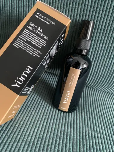 Ένα μαύρο μπουκάλι Room Mist Pure Powder της YUMA δίπλα στη συσκευασία του πάνω σε πράσινο ριγέ ύφασμα.
