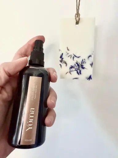 Ένα χέρι κρατά το Fresh LINEN room spray της YUMA δίπλα σε κρεμαστό αρωματικό κερί.