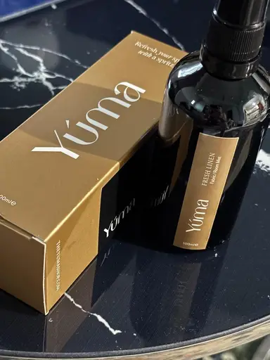Ένα σκουρόχρωμο μπουκάλι room spray Fresh LINEN δίπλα στη χρυσή συσκευασία της YUMA πάνω σε μάρμαρο.