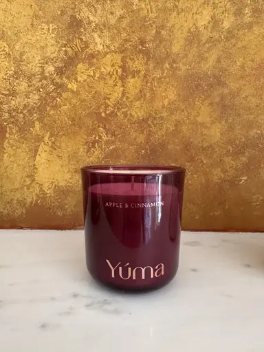 Αρωματικό κερί YUMA Apple & Cinnamon σε μπορντό γυάλινο δοχείο μπροστά από χρυσό τοίχο.
