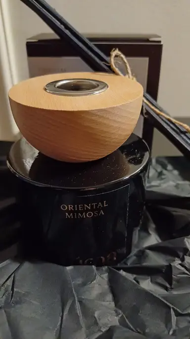 Μαύρο γυάλινο δοχείο Oriental Mimosa με ξύλινο καπάκι και μαύρα στικ αρώματος σε τσαλακωμένο χαρτί.