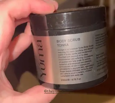 Ένα χέρι κρατάει το Body Scrub TONKA της YUMA σε μαύρο βαζάκι με ασημί ετικέτα.
