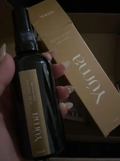Ένα χέρι κρατά το Pure Powder room mist της YUMA δίπλα στη μπεζ συσκευασία του.