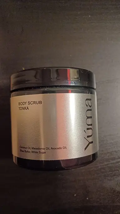 Ένα μαύρο βάζο Body Scrub Tonka της YUMA με ασημί ετικέτα σε σκούρο φόντο.