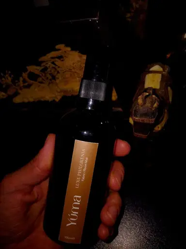 Ένα χέρι κρατά το Luxe Philoxenia Room Mist της YUMA σε σκούρο μπουκάλι με χρυσή ετικέτα.