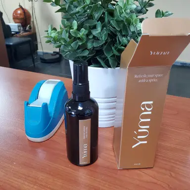 Ένα σκούρο μπουκάλι room spray Yuma δίπλα στη συσκευασία του και ένα μπλε σελοτέιπ.