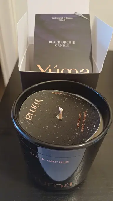 Κερί Black Orchid της YUMA σε μαύρο γυάλινο δοχείο μπροστά από την ανοιχτή συσκευασία του.